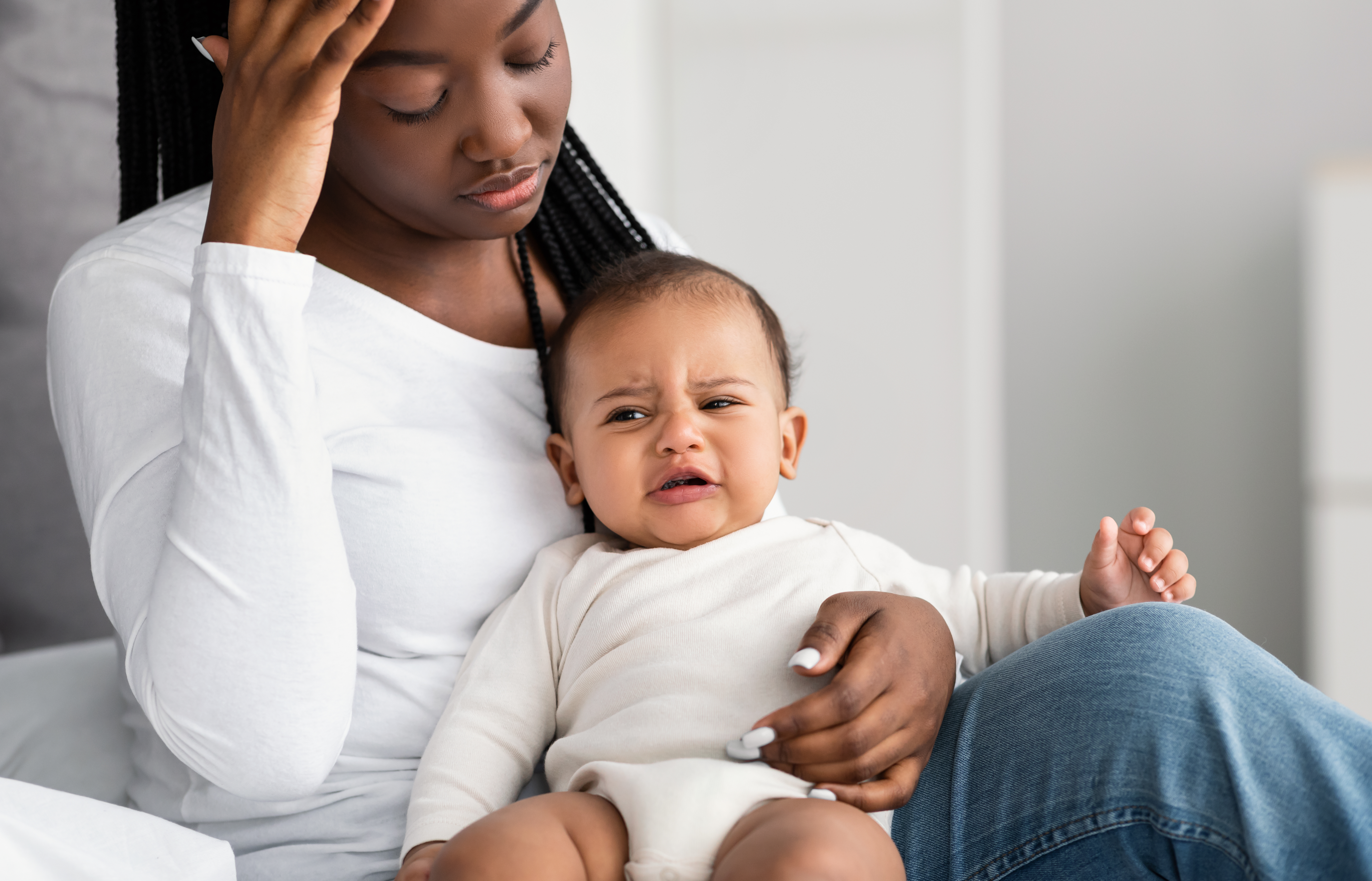Postnatal Stress Concept. Portrait of depressed African American woman sitting with her crying little child on bed at home, feeling ppd baby blues, headache or migraine, touching forehead